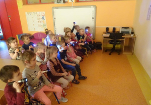 Grupa dzieci siedzi na krzesłach, trzymają włączone latarki skierowane na ścianę.